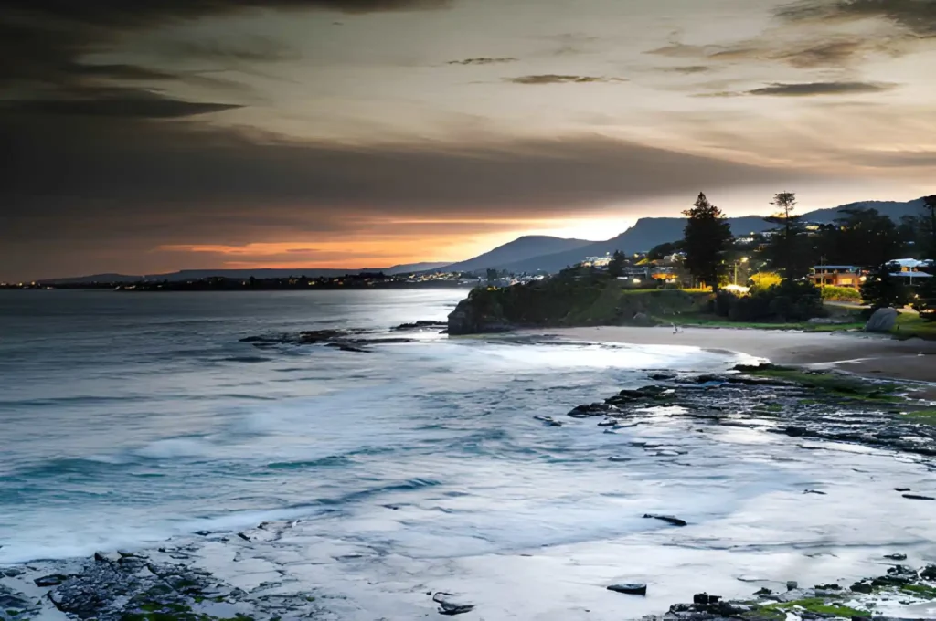 Dramatic sunset over the rocky coast and beach in Farnworth, featuring coastal town lights and distant mountains.
